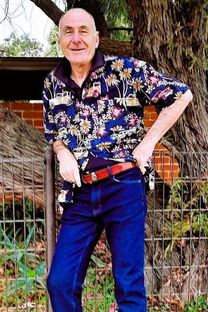 Smiling man wearing a colourful Hawaiian shirt with a blue background. He is also wearing blue jeans.