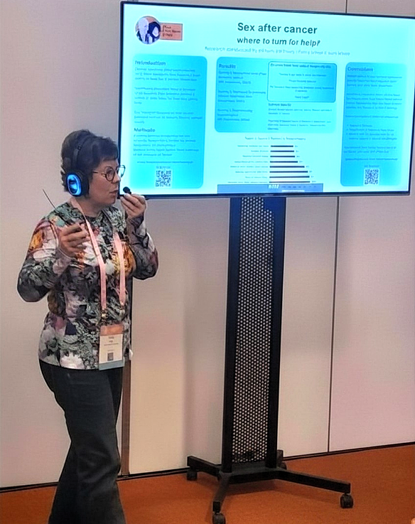 Photograph of a woman wearing headphones and talking into a microphone. Behind her is a video screen that shows her research.