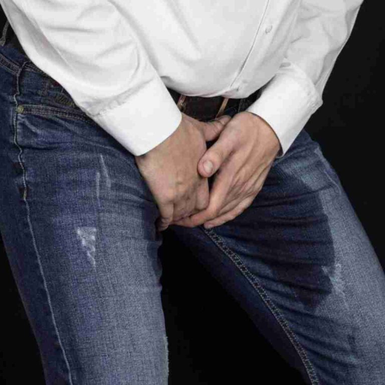 Man in white shirt and jeans clutching his groin with a wet urine patch on his jeans