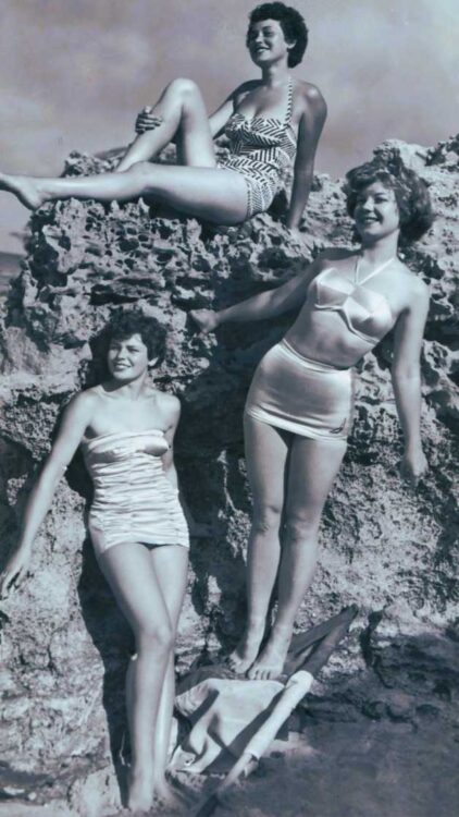 Grayscale photo from 1950s of three young women in bathing suits against a large rock