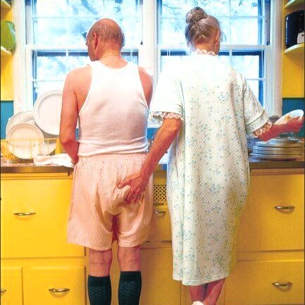 Older couple washing dishes together. Viewed from behind, the woman is giving the man's bottom a squeeze.