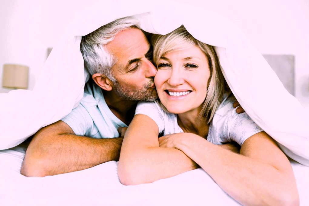 Man with grey hair kissing woman on the cheek as they both lie in bed