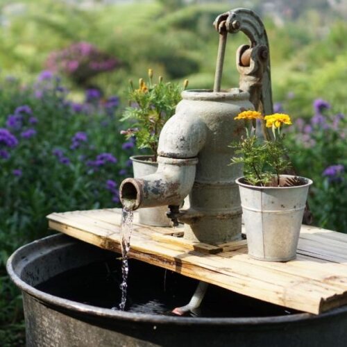 Old-fashioned metal water pump flowing into metal bucket with background of flowers